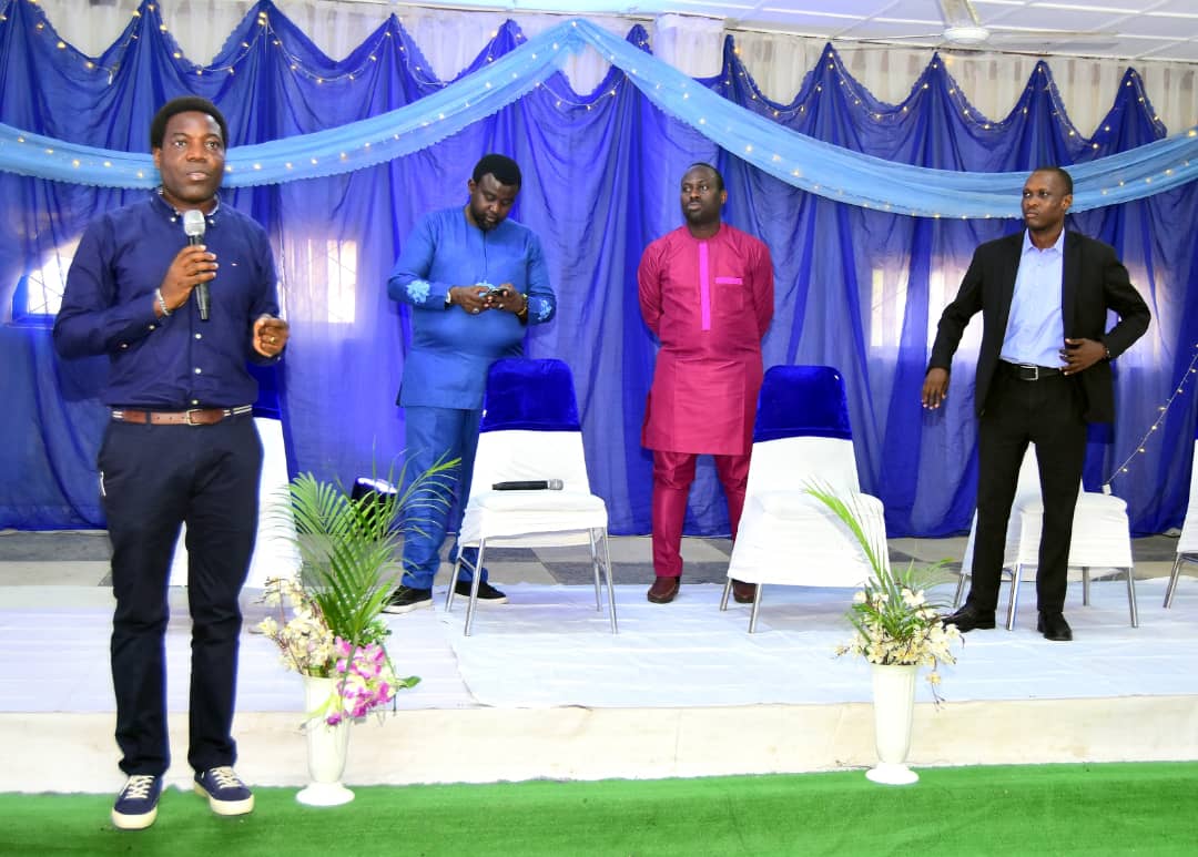 A speaker addressing the room while three contributors stand on the stage